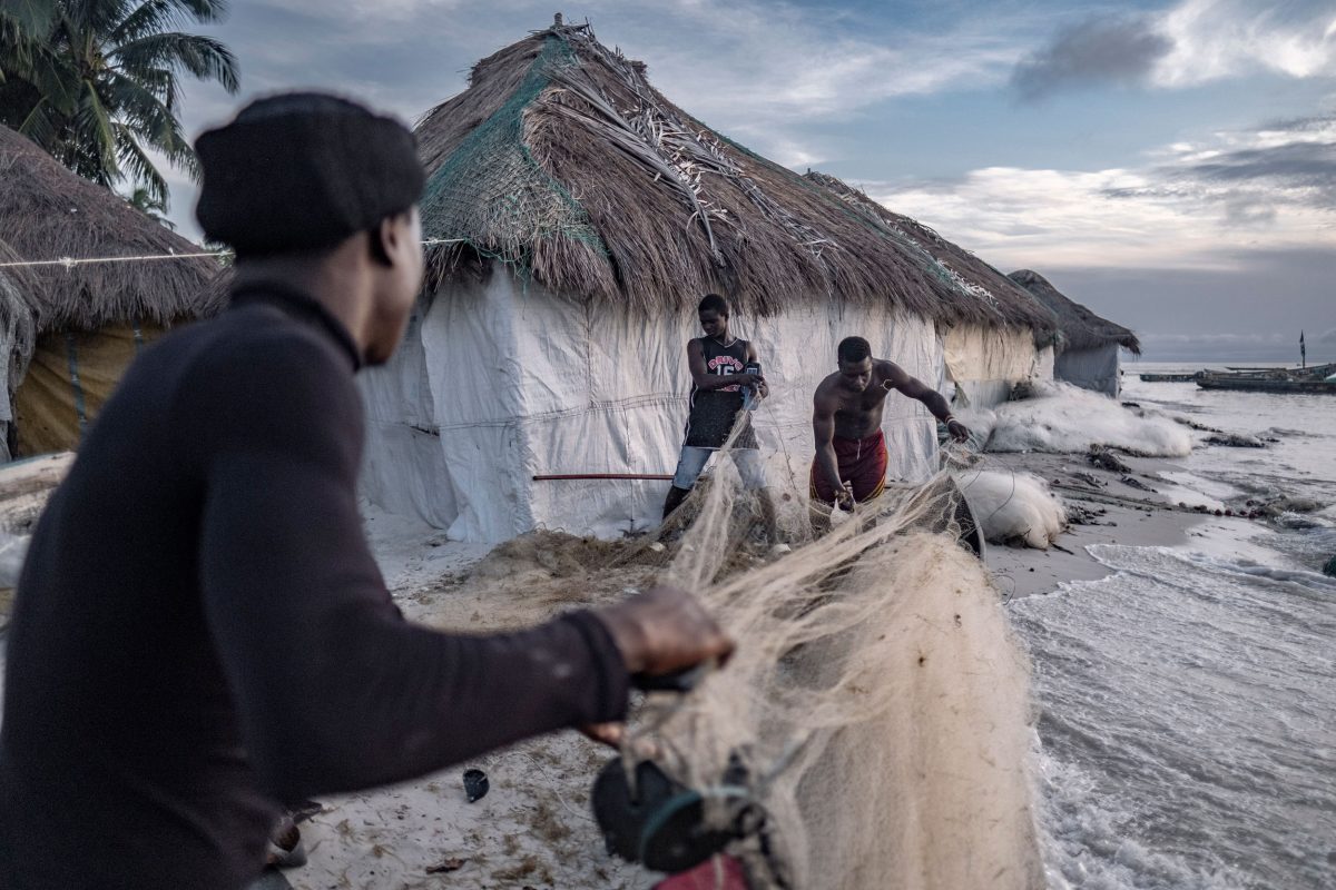Fishermen prepare their nets while a rising tide approaches nearby homes.