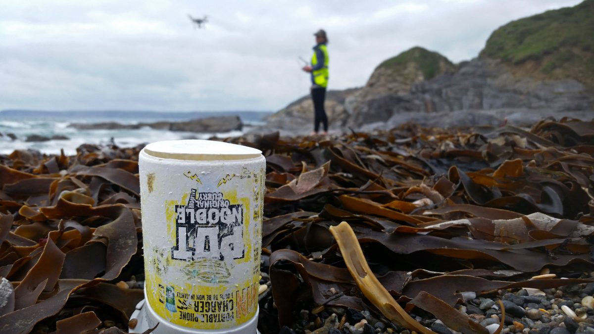 A plastic noodle pot litters Hemmick Beach in Cornwall, England, as Ellie Mackay, cofounder of the Plastic Tide, uses a drone to document plastic debris in the distance. Photo by the Plastic Tide
