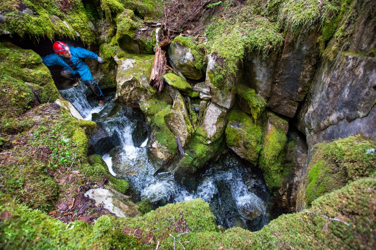 A scientist scrambles over rocky terrain where a river flows out of rock and then back into it.