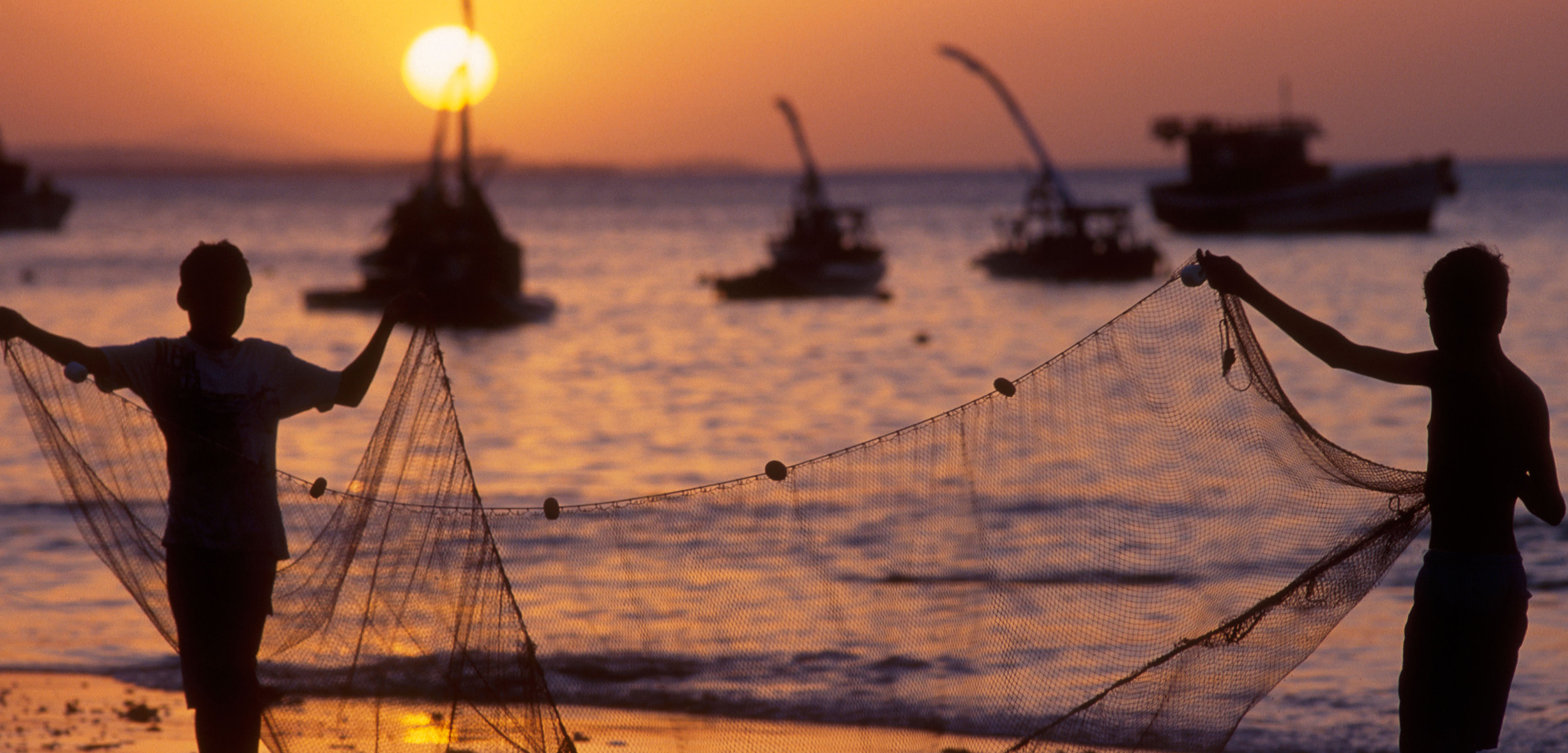 the silhouette of two children holding a fishing net over a sunset with fishing boats in the background