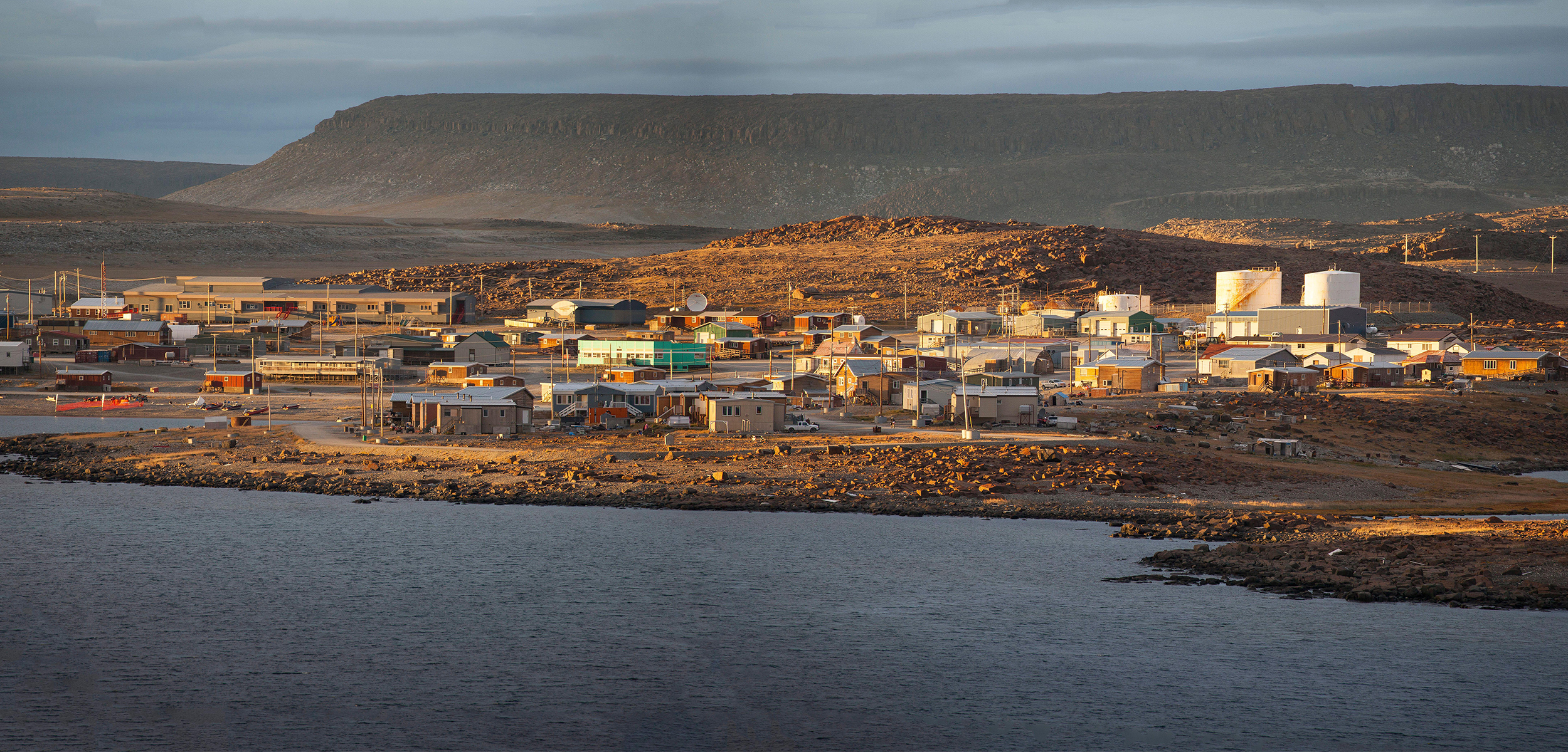A shot from across the water of a small village of homes in a landscape of small brown rollling hills in the tundra.