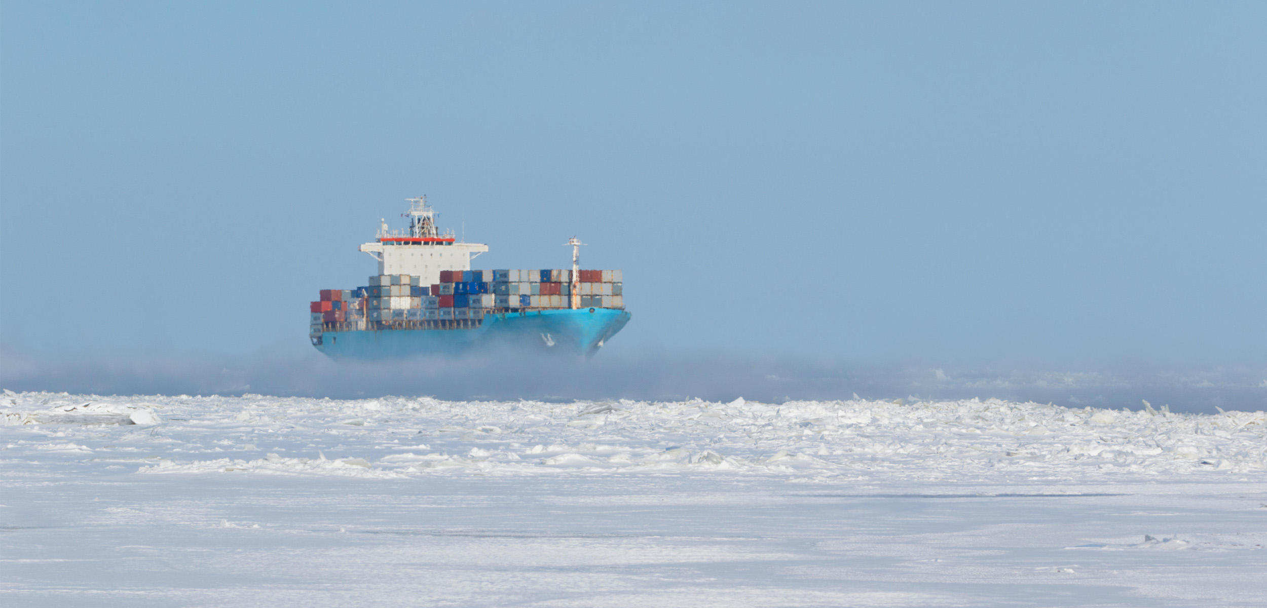 Snow packed ice sitting atop a cold ocean with a blue boat carrying shipping containers