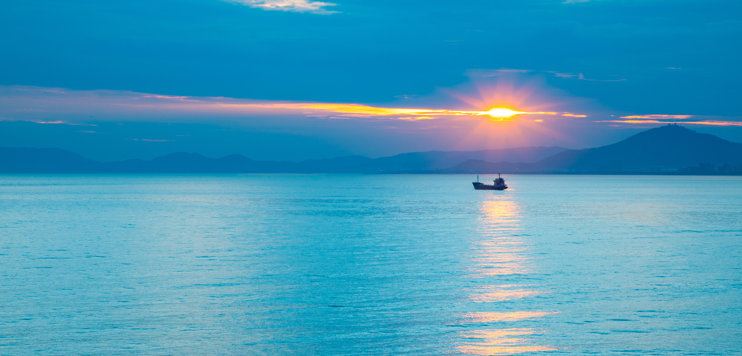 A little boat sitting on the horizon of light blue water with the sun setting behind