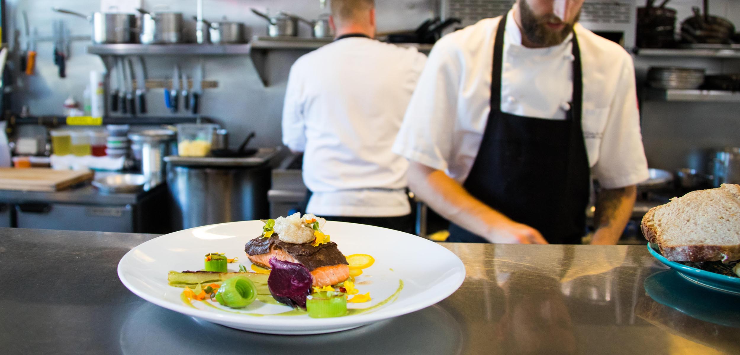 a plate of colourful food sits at the front with the chef cooking it sitting behind wearing a black apron and white shirt
