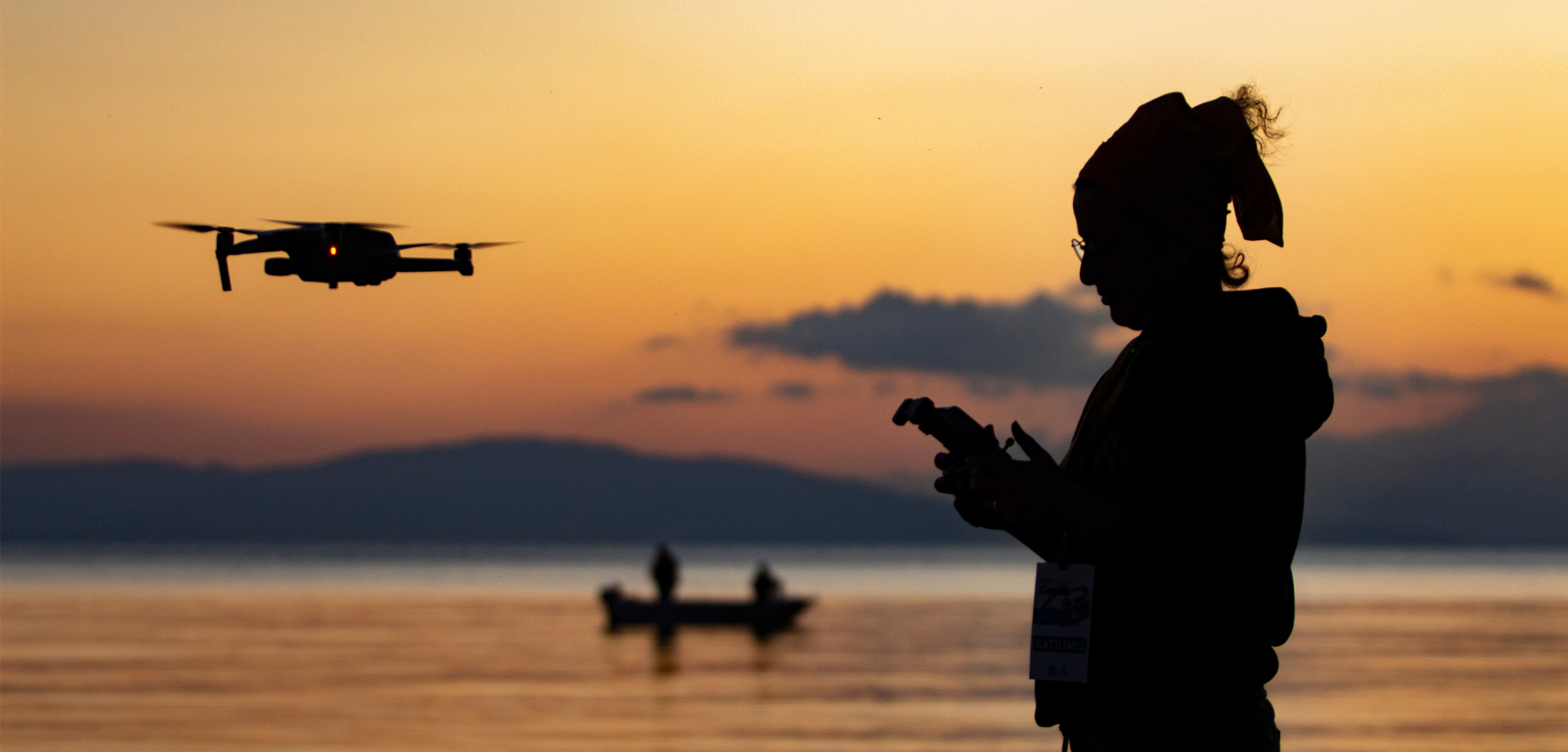 silhouette of young woman flying drone at sunset. Sea and fishing boat in the background. drone and woman taken in reverse light.