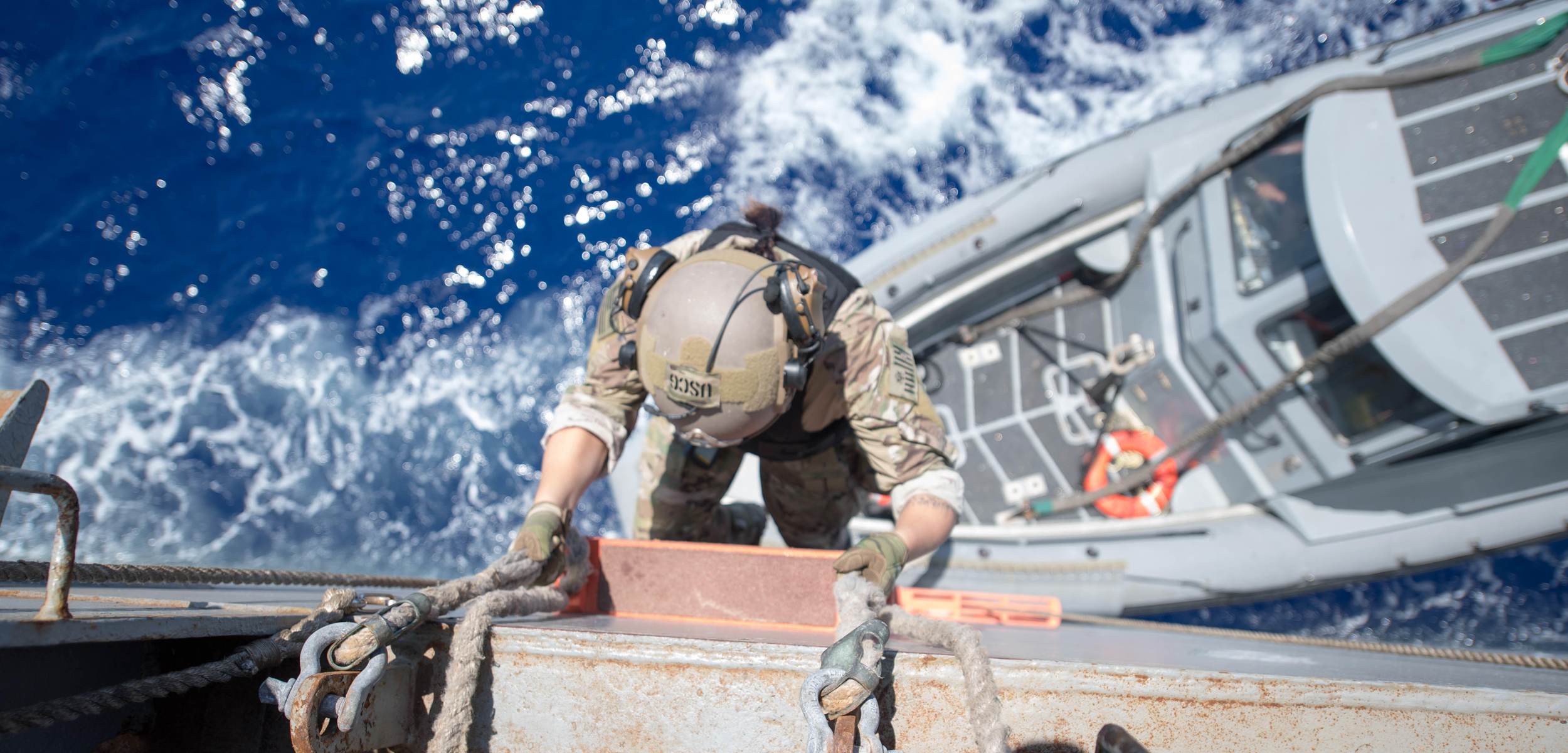 A birds eye view of a coast guard dressde in camoflauge climbing a ladder to board a ship with the blue ocean below.
