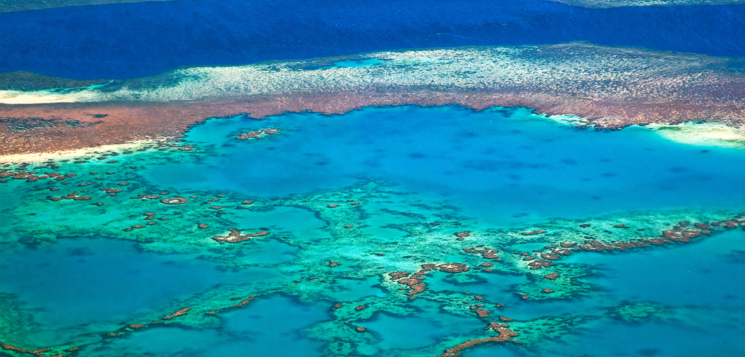 An aerial view of a bright blue ocean dotted with brown and black dots of the reef
