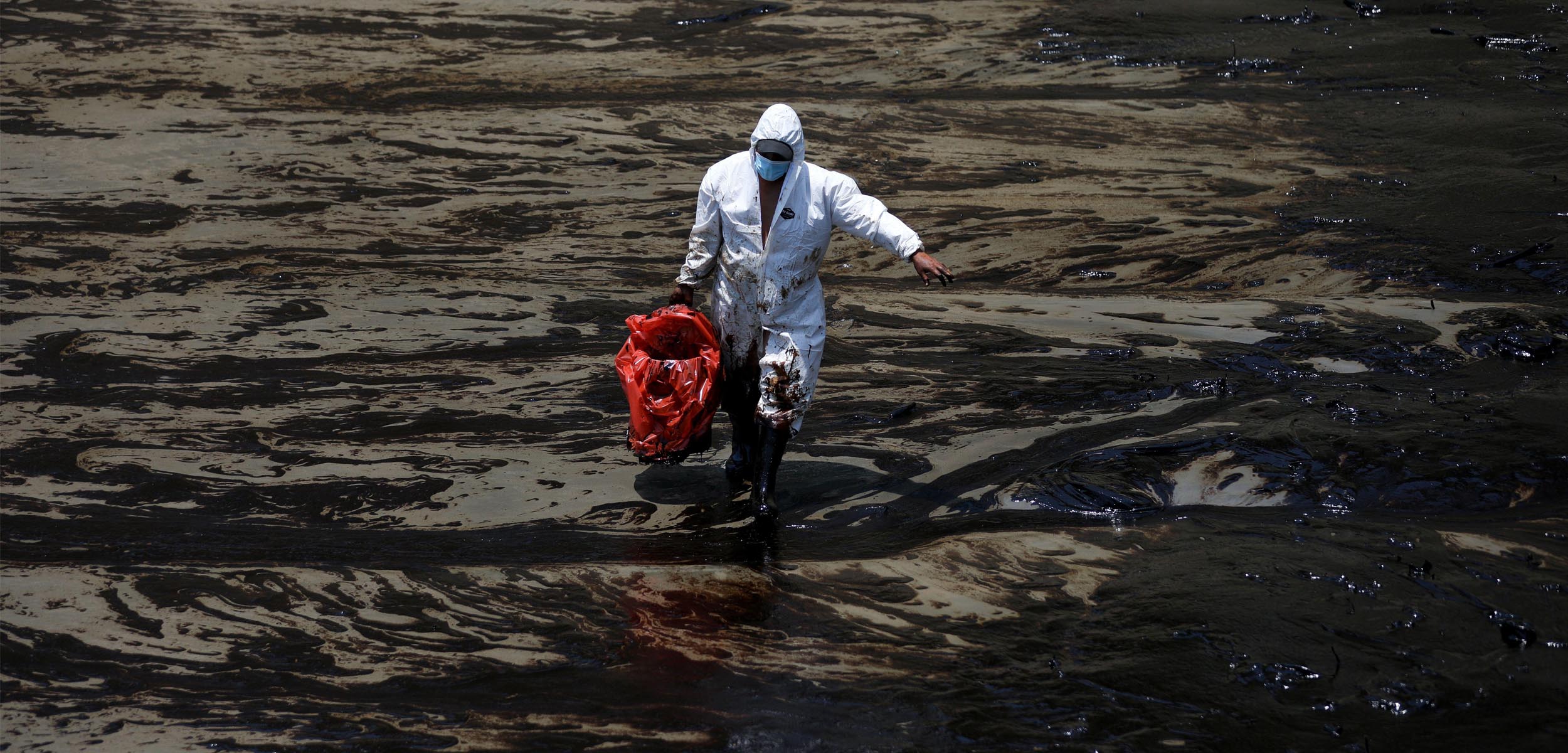 A cleanup worker dressed in a white hazmat suit walks through a black oil covered landscape alone.