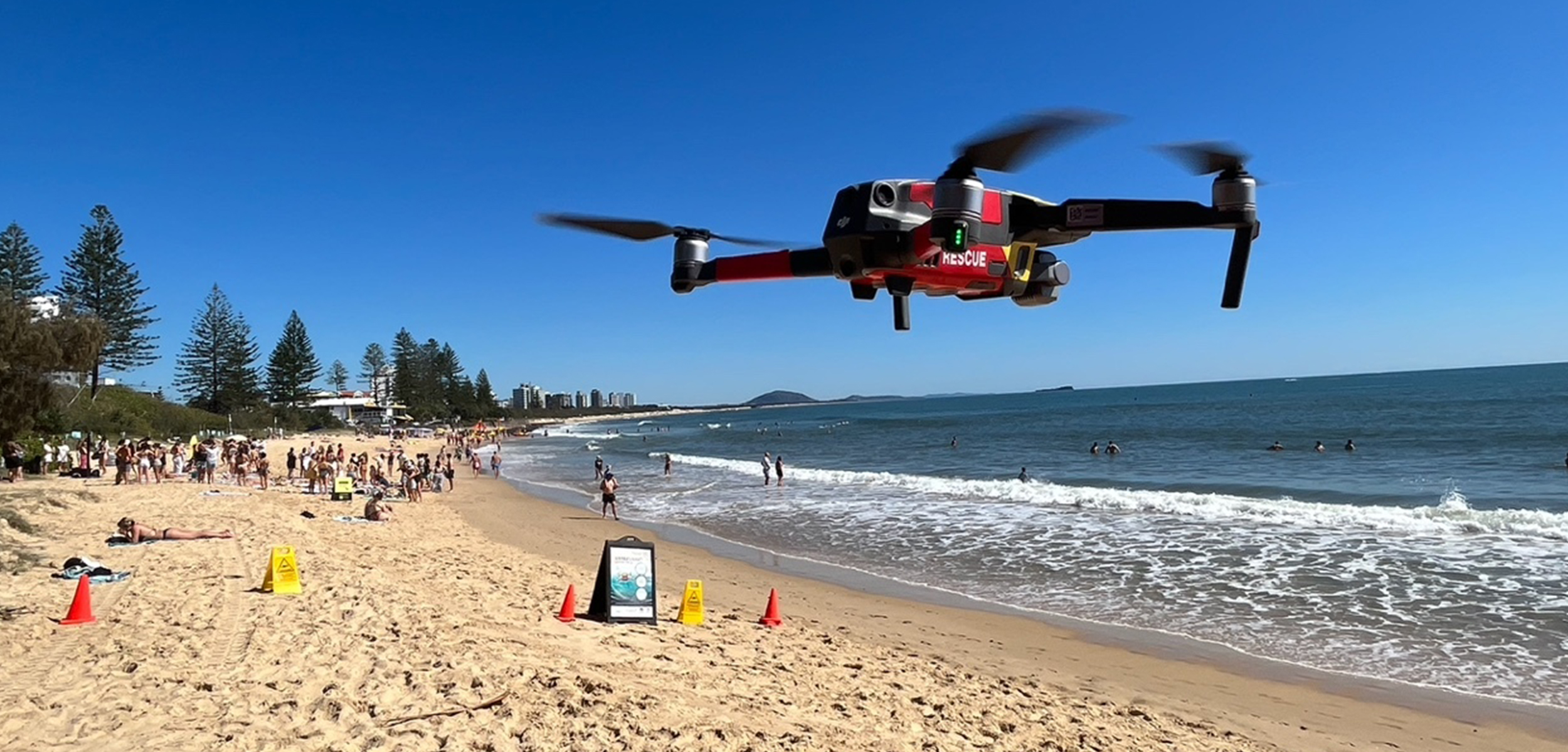 A red drone flys in the blue sky above a yellowy beach with the turquoise ocean below.