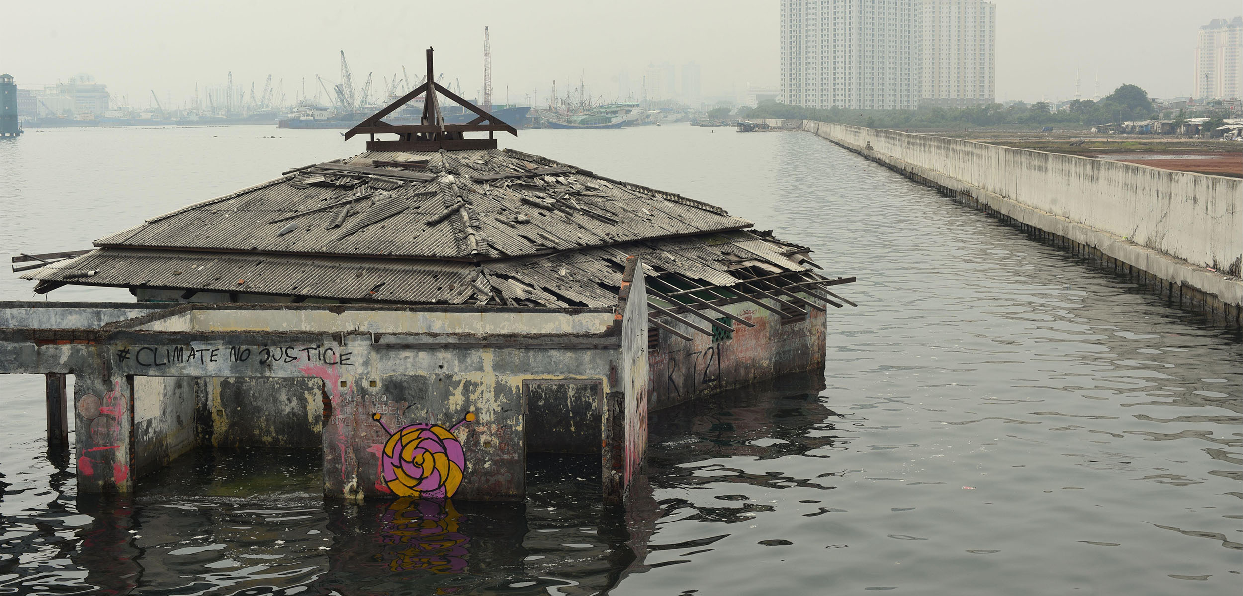 an abandoned building sits falling apart at the front left of the photo with flooding surrounding it and a smog covered city sits in the distances.