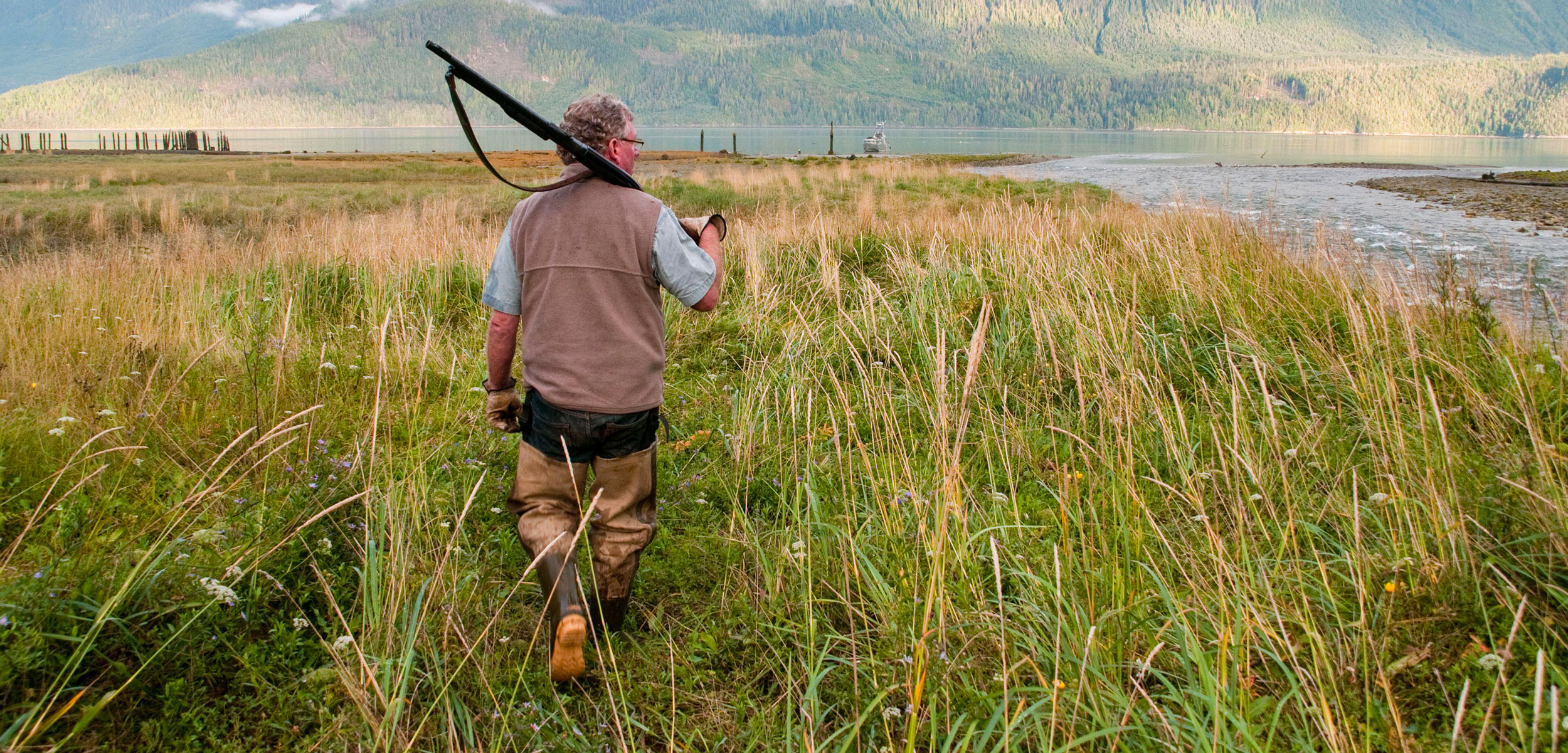 A hunter holding a shotgun and wearing fishing waders in a creek estuary in the Great Bear Rainforest