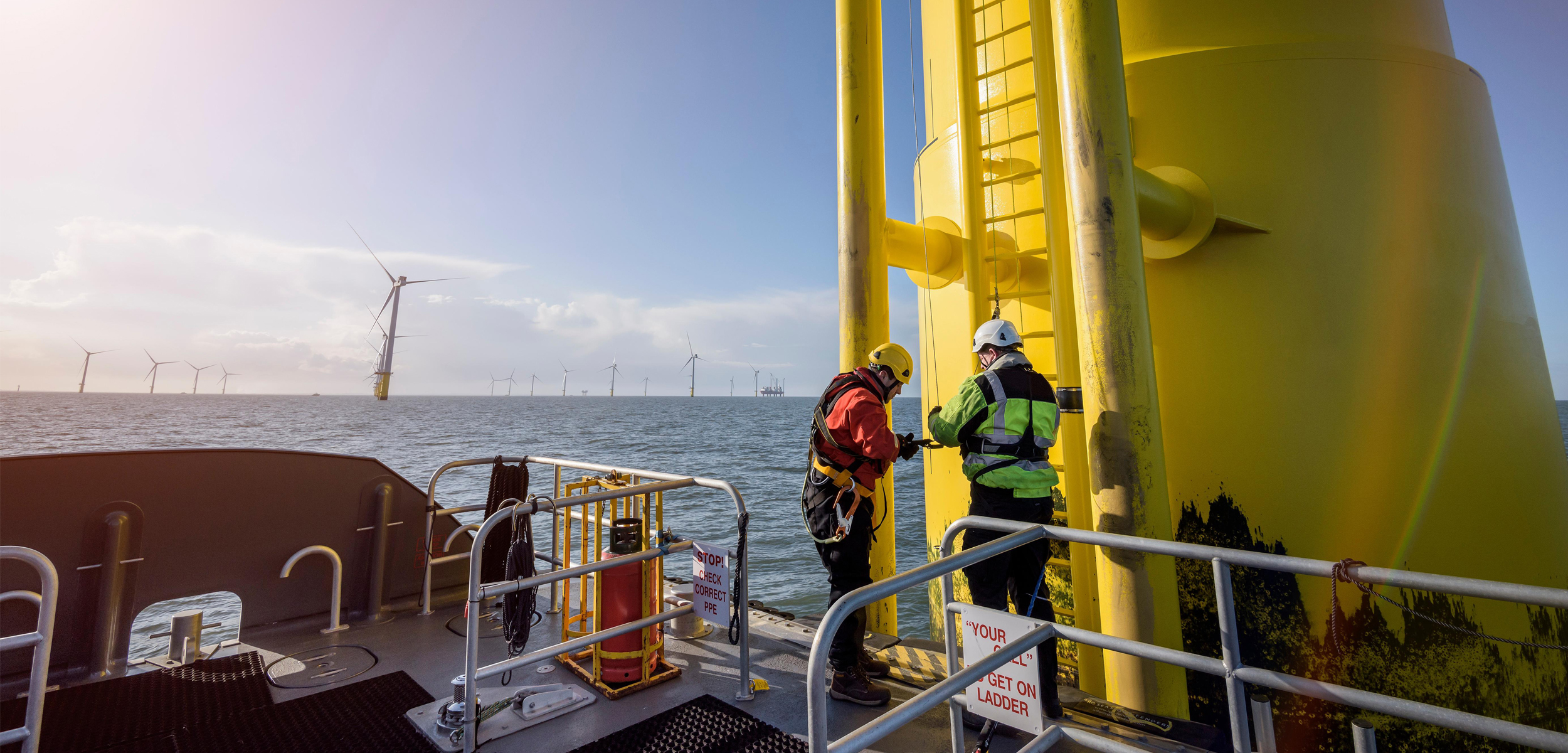 A ship with a large yellow win turbine to the right of the photo. Two men stand in the foreground and in the background you can see many offshore turbines in a blue ocean