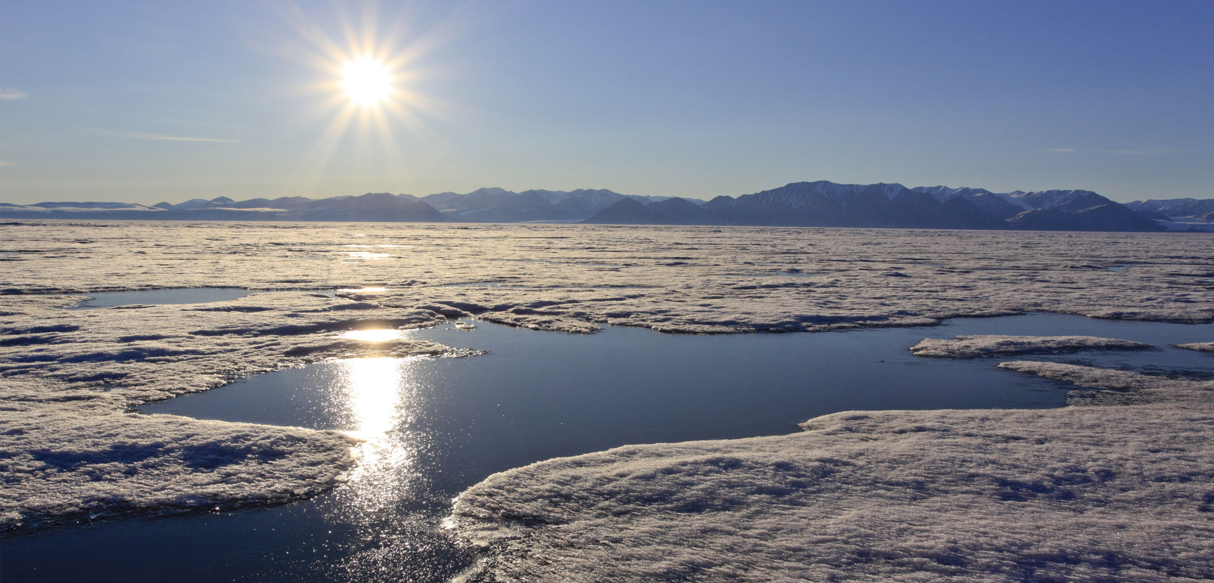 The sun sits above an ice sheet that is has a river flowing through it with mountains through it.