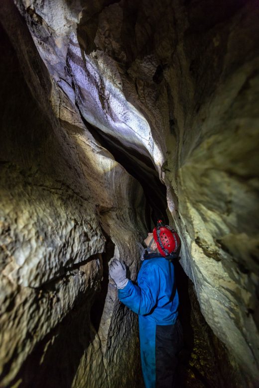 A scientists squeezes through a narrow but tall opening within a cave.